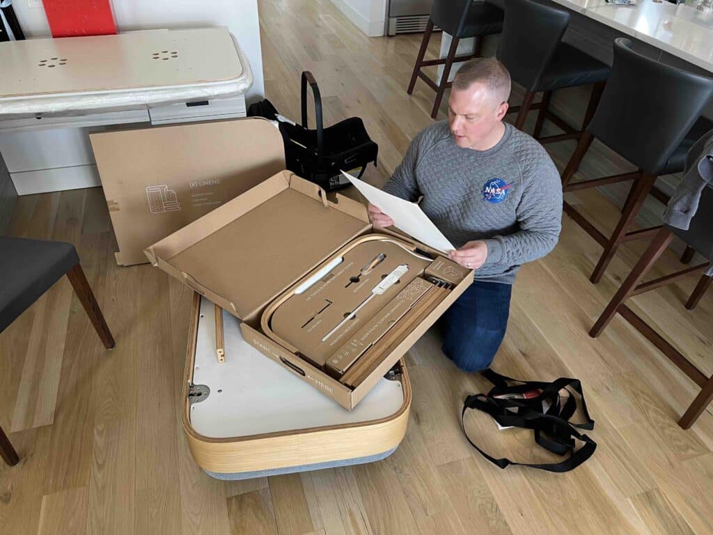 A man starts unboxing Cradlewise to reveal a set of provided tools. He is kneeling on the floor, wearing a NASA sweatshirt, and reviewing an instruction booklet