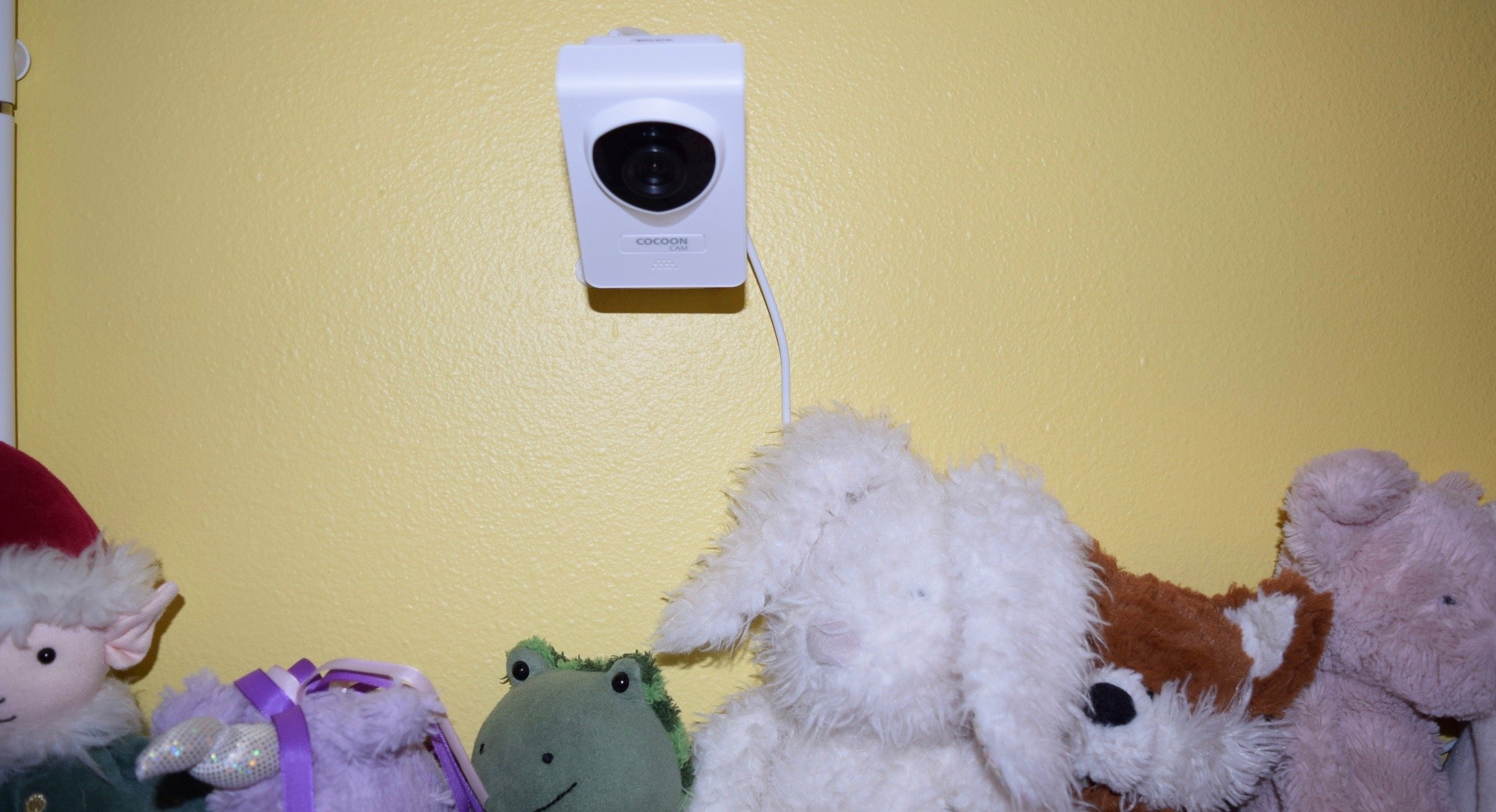 A Cocoon Cam mounted on a yellow wall above a crib with stuffed animals on the railing