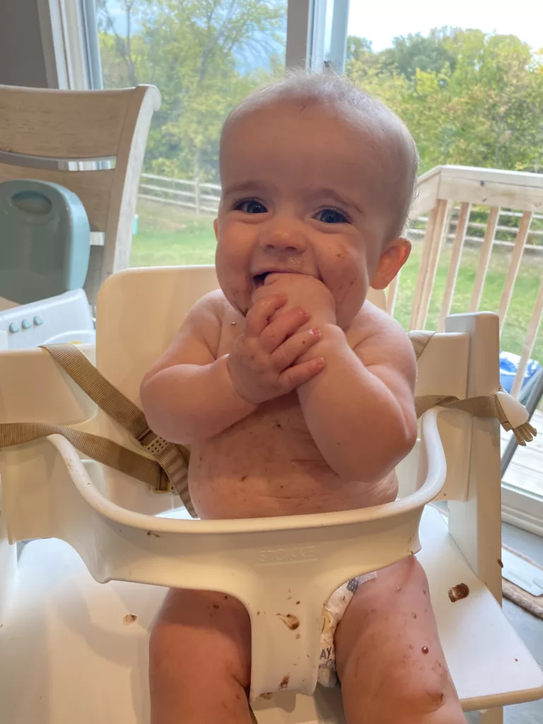 This is a close-up image of an infant sitting in the Stokke high chair. He is covered in a puree, eating, and smiling at the camera.