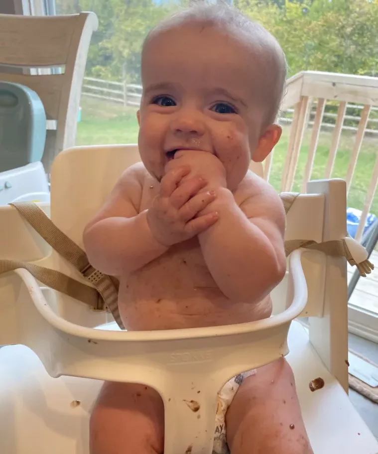 This is a close-up image of an infant sitting in the Stokke high chair. He is covered in a puree, eating, and smiling at the camera.