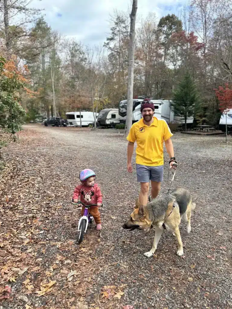 A man (the author), a German Shepard, and a toddler are together on a gravel road at a campsite, while the toddler rides a bike