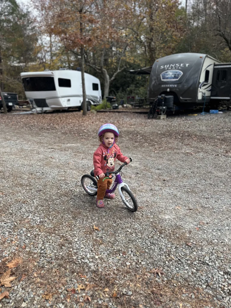 A toddler rides the Woom 1 on a gravel road. She is at a campsite, with camper vans in the background. 