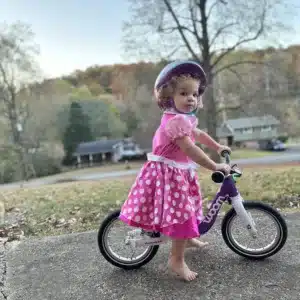 A toddler in a pink dress rides the Woom 1 on a sidewalk. She is wearing a helmet and looking at the camera.
