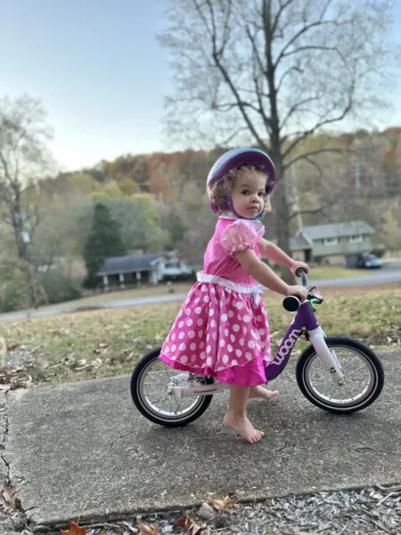 A toddler in a pink dress rides the Woom 1 on a sidewalk. She is wearing a helmet and looking at the camera.